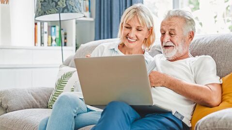 Mann und Frau sitzen lachend auf einem Sofa und schauen auf einen Laptop