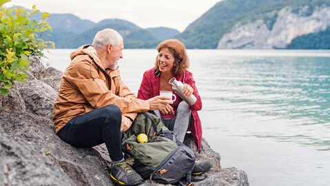 Frau und Mann sitzen an einem See in den Bergen, trinken Tee und essen einen Apfel