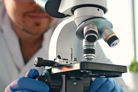 Mann mit weißem Kittel und blauen Hygienehandschuhen bedient ein Mikroskop