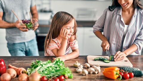 Frau schneidet eine Gurke, während ihre Tochter dabei zuschaut. Im Hintergrund steht ein Mann mit einer Salatschüssel in der Hand. Im Vordergrund liegen Karotten, Paprika, Pilze, Tomaten und Salat.