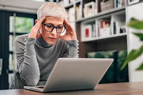 Frau mit kurzen Haaren und Brille sitzt vor einem Laptop und hält sich mit beiden Händen an der Stirn