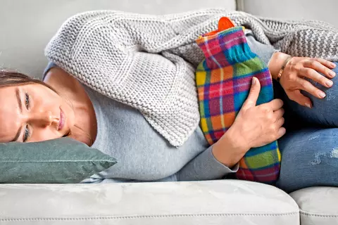 Frau liegt mit starrem Blick auf dem Sofa und drückt eine bunte Wärmeflasche auf ihren Bauch