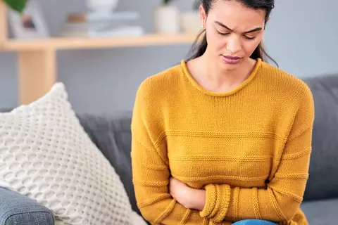 Frau mit gelbem Oberteil sitzt gekrümmt auf der Couch und hält sich den Bauch aufgrund anhaltender Blähungen