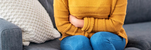 Frau mit gelbem Oberteil sitzt gekrümmt auf der Couch und hält sich den Bauch aufgrund anhaltender Blähungen