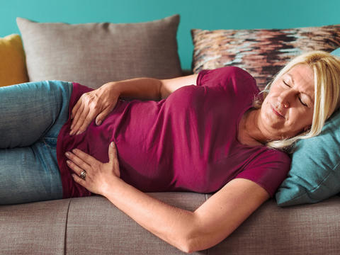 Frau liegt mit geschlossenen Augen auf der Couch und hält sich am Bauch