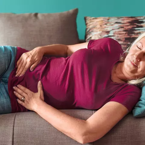 Frau liegt mit geschlossenen Augen auf der Couch und hält sich am Bauch