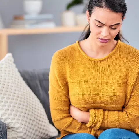 Frau mit gelbem Oberteil sitzt gekrümmt auf der Couch und hält sich den Bauch aufgrund anhaltender Blähungen